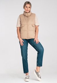 Beżowa kamizelka puchowa z wysokim kołnierzem, z dużymi przednimi kieszeniami, zestawiona z jasnoniebieskimi dżinsami i sneakersami w zwierzęcy wzór.