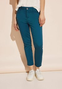 Tyrkysové elastické chino kalhoty s přiléhavým střihem, vybavené předními kapsami, zapínáním na jeden knoflík a srolovanými lemy. Kombinovány s bílými teniskami.