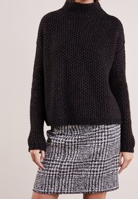 Schwarzer, strukturierter Strickpullover mit hohem Kragen, kombiniert mit einem figurbetonten Rock in schwarz-weißer Hahnentrittmusterung. Der Pullover hat überschnittene Schultern.