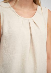 Beige ärmelloses Oberteil mit rundem Ausschnitt und subtilen Falten an der Vorderseite, aus strukturiertem Stoff gefertigt. Einfaches Design und neutrale Farbe.