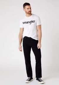 Maglietta bianca con logo "Wrangler", jeans neri e sneakers nere. L'outfit presenta una vestibilità comoda con un design classico.