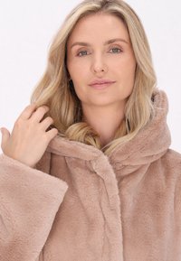 Cappotto con cappuccio in finta pelliccia di un morbido beige, con maniche ampie e una texture soffice, dotato di chiusura frontale e una vestibilità rilassata.