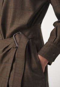 Robe à carreaux marron avec manches longues, poignets boutonnés, et ceinture en tissu assorti avec une boucle en métal attachée à la taille.