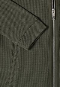 Olivengrøn ribstrikket jakke med lange manchetærmer og sølvfarvet metallisk lynlås, der løber lodret.