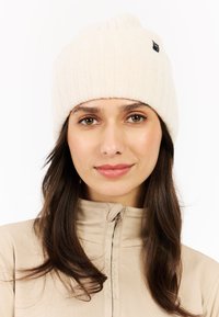 Cream knitted beanie with ribbed texture, featuring a small black tag on the side. Worn with a light-colored pullover.
