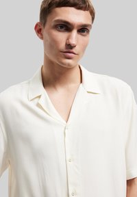 Camisa corta de manga en color crema, confeccionada en una tela ligera, con cuello notch y cierre de botones, que presenta una textura suave y un sutil brillo.