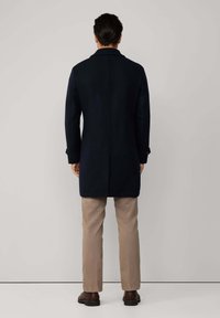 Uomo di spalle che indossa un lungo cappotto blu scuro, pantaloni beige e scarpe marroni, in piedi contro uno sfondo grigio chiaro uniforme.