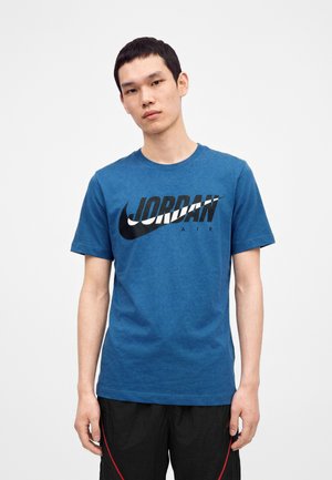 Ung mand iført en blå Jordan Air t-shirt og sorte bukser, stående foran en ensfarvet lys baggrund.