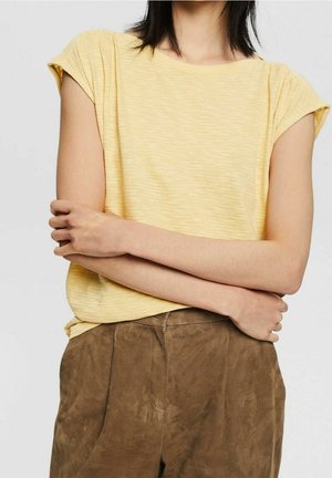 Femme aux bras croisés portant un haut à manches courtes jaune pâle et un pantalon ample marron, sur un fond uni.