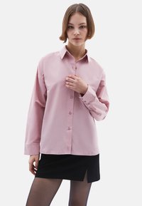Lichtroze langarm blouse met knoopsluiting en kraag, met een gladde textuur en een relaxte pasvorm. G gedragen met een zwarte rok, gedeeltelijk zichtbaar.