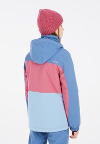 Farbenfrohe, mittelange Jacke in Blau, Pink und Hellblau; verfügt über eine Kapuze und elastische Bündchen. Strickmütze in tiefem Pink.