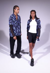 Due persone si trovano su uno sfondo semplice. L'uomo indossa una camicia a maniche corte blu e nera con pantaloni neri. La donna ha una maglietta bianca e pantaloni corti neri, con una camicia fantasia appoggiata su di lei. Entrambi indossano scarpe nere.