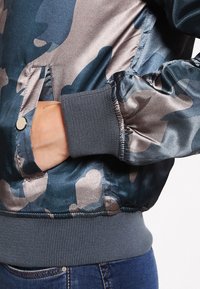 Bomberka w kamuflażowym wzorze, wykonana z satinowego materiału, w ciemnoniebieskim i brązowym kolorze, z prążkowanymi mankietami oraz bocznymi kieszeniami z zapięciem na zatrzask.