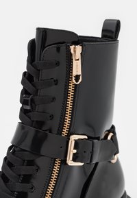 Bottine en cuir verni noir avec fermeture éclair latérale à ferrures dorées, devant à lacets, et une bride à boucle autour de la cheville.