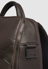 Bruine leren rugzak met een gladde textuur, voorzien van een zichtbare rits aan de zijkant en een metalen merklabel aan de voorkant.