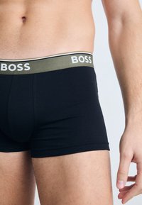 Schwarze Boxerunterhosen mit einem olivgrünen Bund, der die weiße "BOSS"-Markierung trägt. Der Stoff erscheint glatt und hat einen engen Sitz.