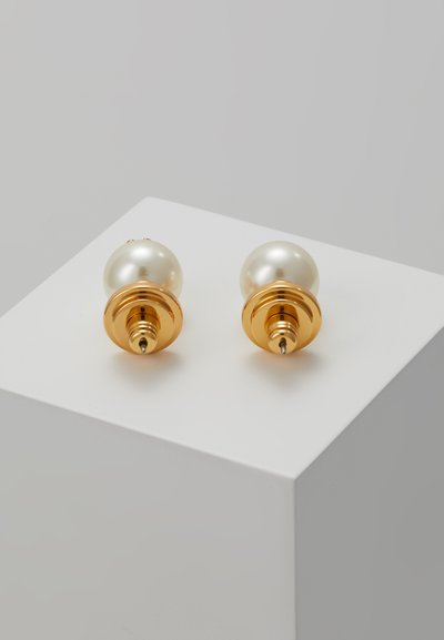 Orecchini a bottone in oro con grandi perle crema lucenti e punte appuntite, adagiati su una superficie bianca di esposizione.