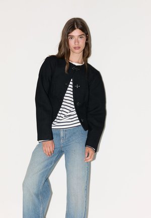 Jeune femme portant une veste noire sur une chemise rayée blanche et noire associée à un jean bleu clair, debout devant un fond blanc uni.