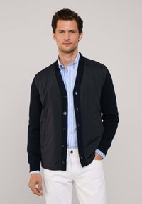 Cárdigan acolchado navy con botones al frente, mangas oscuras en contraste, puños acanalados, combinado con una camisa a rayas azul claro y pantalones blancos.