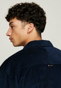 Chemise en velours côtelé bleu marine avec une finition texturée, dotée d'un col et d'une étiquette à l'arrière. Le tissu présente des détails en côtes verticales.