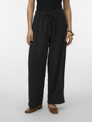 Mujer con pantalones negros sueltos y texturizados con cordón, zapatos marrones de punta cuadrada y una pulsera en su muñeca derecha, de pie contra un fondo blanco.