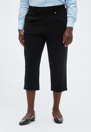 Osoba ubrana w czarne, krótko przycięte spodnie z guzikami, w połączeniu z czarnymi lakierowanymi butami na niskim obcasie typu slingback.