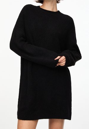 Personne portant une longue robe pull noire en maille côtelée avec de longues manches couvrant une partie des mains, sur un fond blanc uni.