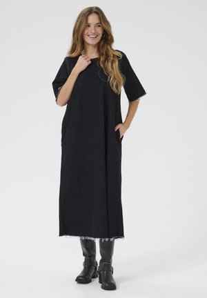 Abito lungo in denim nero con maniche corte, orlo sfilacciato e tasche laterali; indossato con stivali neri fino al polpaccio. Design semplice, vestibilità comoda.