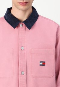 Rosa skjorte med oppkneppede ermer og en kordfløyelsnavi krage, metallknapper og en brystlomme med en liten logo-patch.