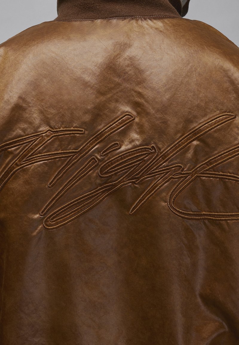 Braune Lederjacke mit glatter Textur, versehen mit großen, erhabenen Buchstaben "Flight" auf dem Rücken, ergänzt durch einen braunen Kragen.