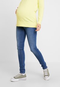 Top amarelo de mangas longas para gravidez combinado com jeans skinnies azuis e ténis cinzas. O conjunto apresenta um corte descontraído e uma textura de tecido suave.