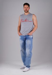Mann steht mit den Händen in den Taschen, trägt ein graues ärmelloses Shirt, helle blaue Jeans und weiße Sneaker vor einem einfarbigen grauen Hintergrund.