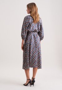 Langärmliges Kleid mit taillierter Passform, gemustert mit kreisförmigen Motiven in Blau, Rot und Creme. Weicher Stoff, Midi-Länge, kombiniert mit schwarzen Absätzen.