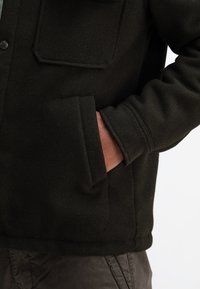 Black fleece jacket with a structured design, featuring a chest pocket and side pockets, and a soft texture.