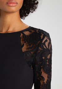 Czarna, długorękawowa odzież z gładką górną częścią i misternymi czarnymi koronkowymi rękawami, które prezentują kwiatowe wzory i gładką fakturę.