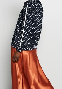 Giacca navy a pois con strisce bianche sulle maniche abbinata a una gonna in satin arancione, che mette in risalto una trama liscia e un design fluido.