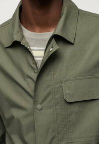 Giacca leggera verde oliva con colletto a camicia, tasca frontale e chiusura a scatto; indossata sopra un maglione a righe. Texture morbida, design casual.