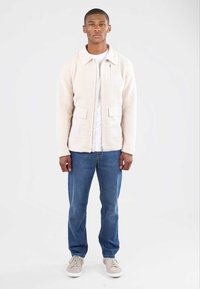 Cremfarbener Fleece-Jacke mit einem Kragen, zwei vorderen Taschen und einer Reißverschlusstasche auf der Brust. Getragen über einem weißen T-Shirt und kombiniert mit blauen Jeans.