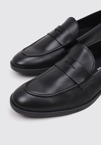 Mocasines de cuero negro con una textura suave, que presentan un diseño de cinta simple y sutiles detalles de costura a lo largo de los bordes.