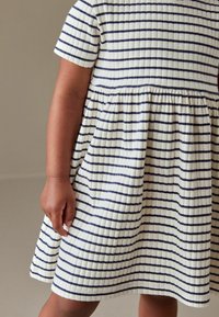 Biała sukienka w granatowe paski, krótki rękaw i marszczony pas. Materiał ma teksturowany, prążkowany wygląd.