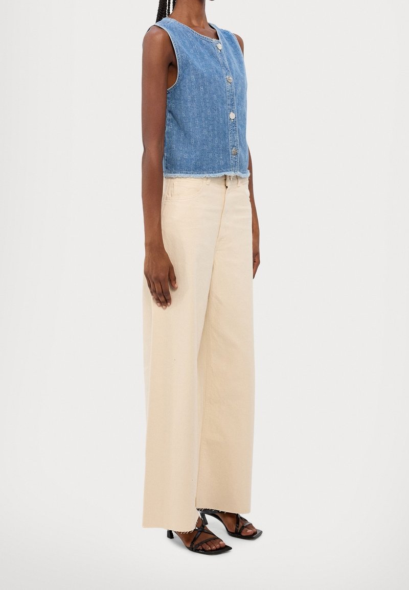 Top sin mangas de denim con botones al frente y dobladillo deshilachado; combinado con pantalones beige de pierna ancha y sandalias negras con tiras. Fondo blanco liso.