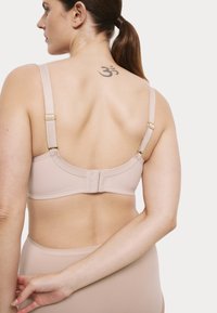 Beige bh met verstelbare bandjes en gouden afwerkingen. Glad stof met een zachte textuur en een haak-en-oogsluiting. Zichtbare tatoeage op de rug.