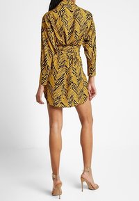 Frau in einem gelb-schwarzen Tigerprint-Minikleid mit langen Ärmeln und passendem Gürtel, kombiniert mit hautfarbenen High-Heels, stehend.