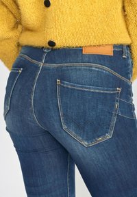 Denim jeans in dark wash with orange stitching and a leather label at the waistband. Features two back pockets with distinct curved designs.