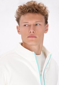 Witte zip-up jas met een hoge kraag en turquoise ritsaccenten, gedragen over een bijpassend wit shirt. Glad stof met een zachte textuur.