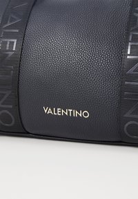 Navy leather handbag featuring textured surface, gold "VALENTINO" logo, and black patterned strap with logo detail.