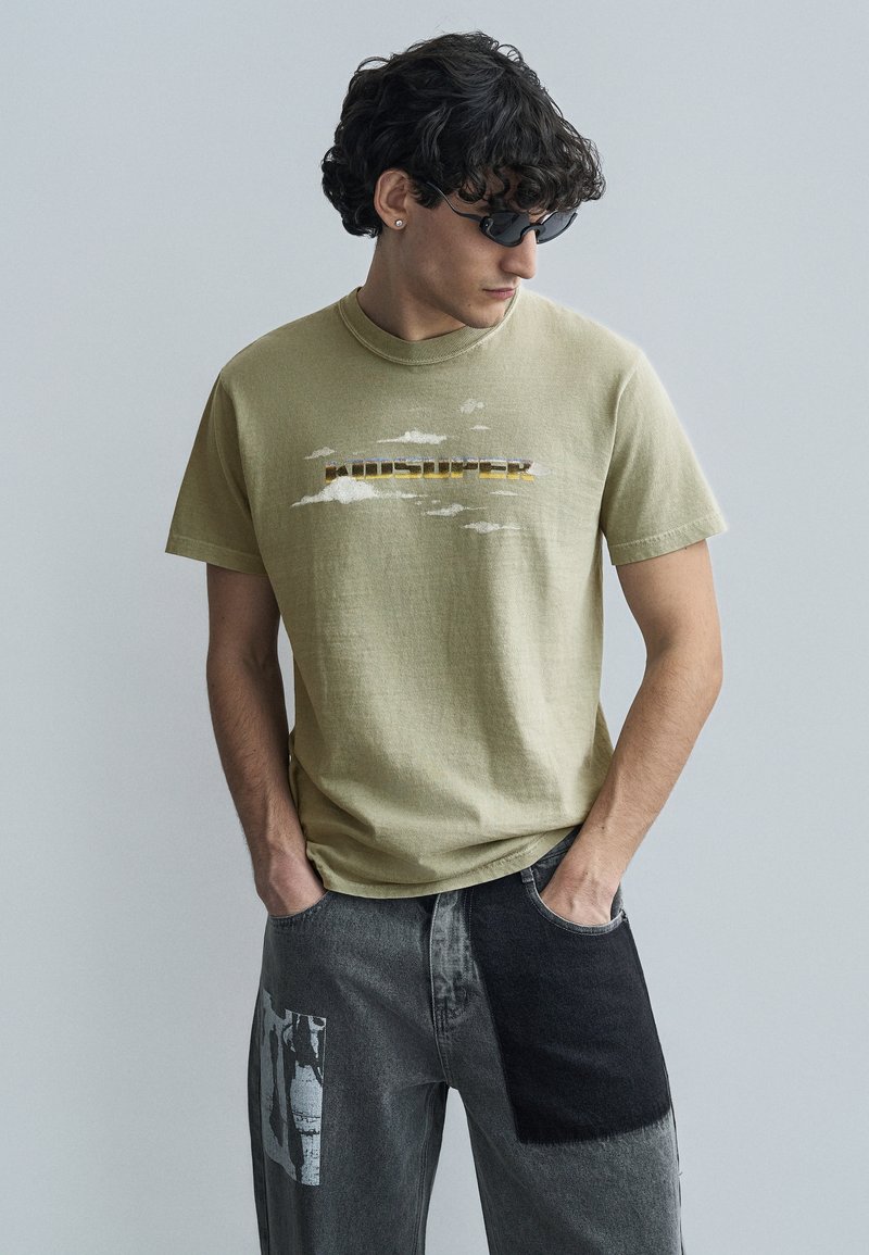 Ανοιχτό πράσινο βαμβακερό t-shirt με σχέδια νεφών και κείμενο "Houdsuper" σε κίτρινο και καφέ. Συνδυασμένο με γκρι τζιν με ανάμεικτες υφές.