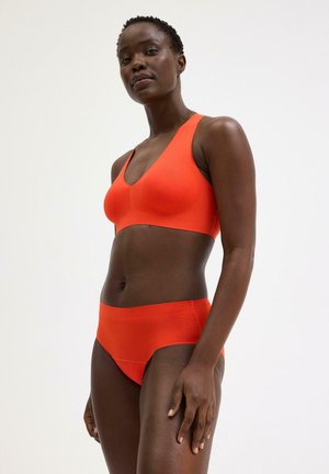 Frau steht und trägt einen leuchtend orangefarbenen nahtlosen BH und hoch taillierte Slips vor einfachem weißem Hintergrund.