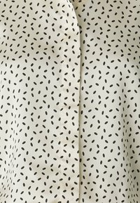 Krémová blúzka s predným švom, s rozptýleným čiernym oválnym vzorom, hladkou textúrou a ľahkým materiálom.