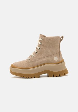 Bottes cheville en toile beige avec une semelle plateforme épaisse en caoutchouc beige, des lacets visibles et un petit logo sur le côté. Tissu texturé.
